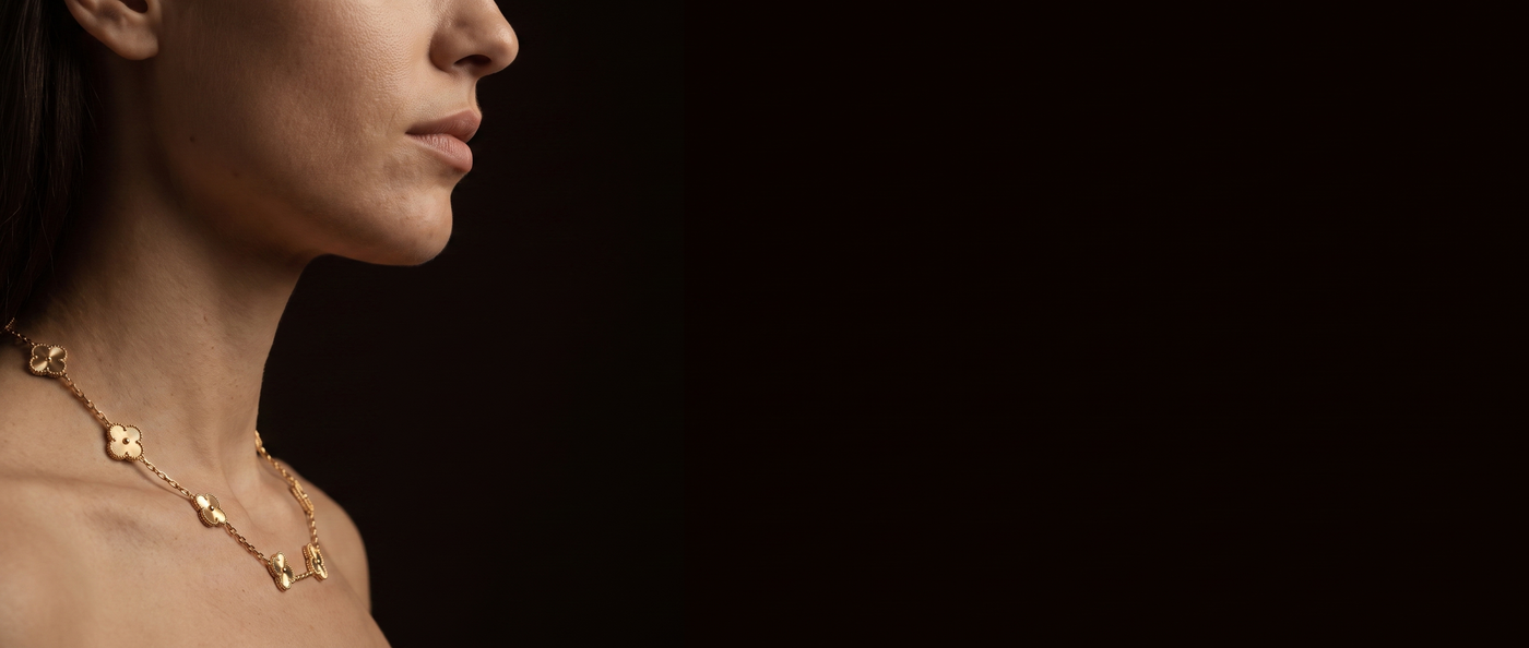 A woman in profile wearing a gold-plated station necklace against a dark background